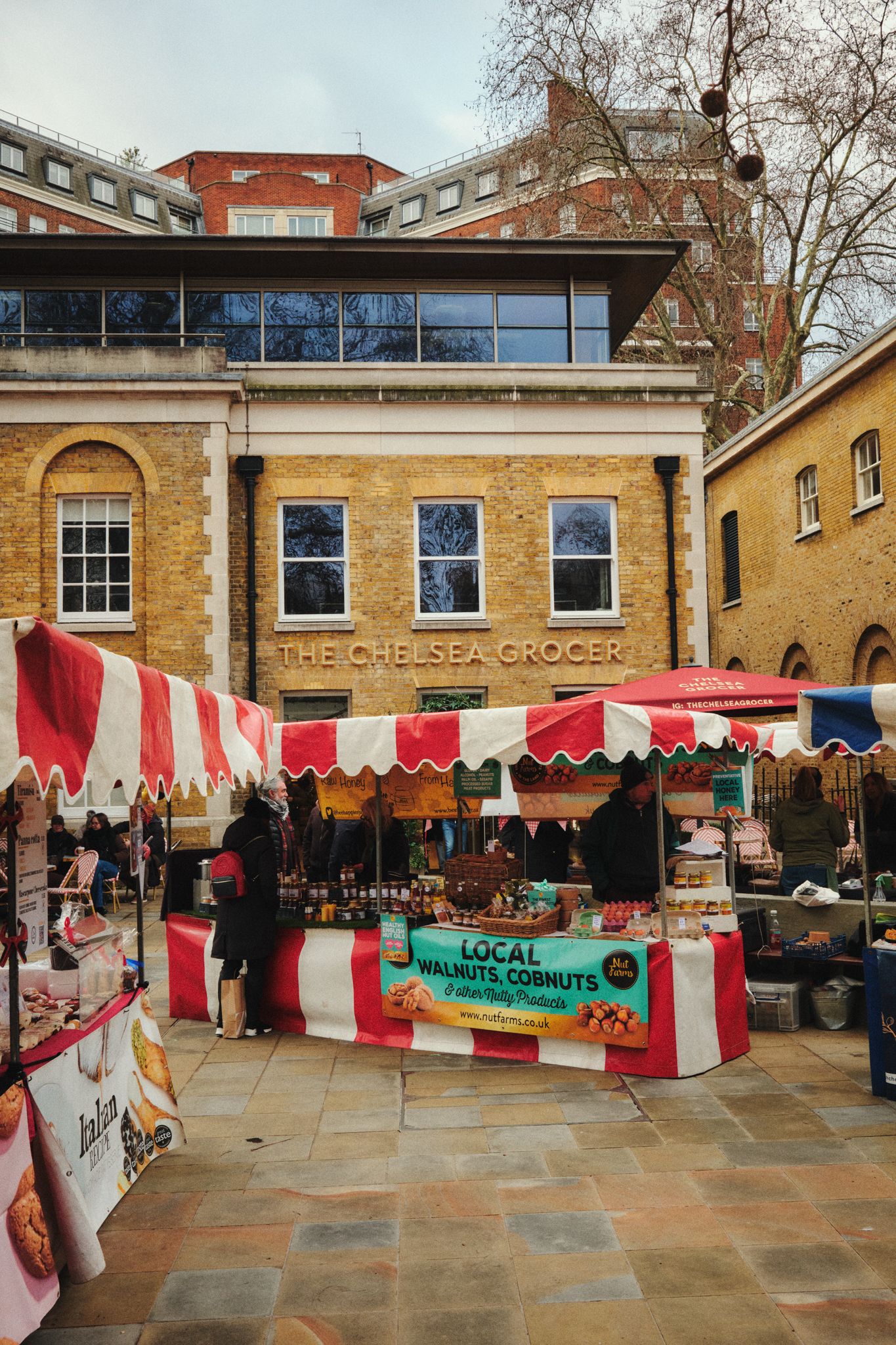 Outdoor food market stalls with red‑and‑white striped canopies set up in a paved square in front of a brick building with a sign reading ‘The Chelsea Grocer’. Sellers display local produce like nuts and other goods, and people browse the market in a relaxed urban setting.