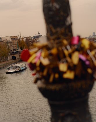 View of a bridge in Paris covered with padlocks, with the Seine River in the background, a boat on the water, and Parisian buildings in the distance under a cloudy sky.
