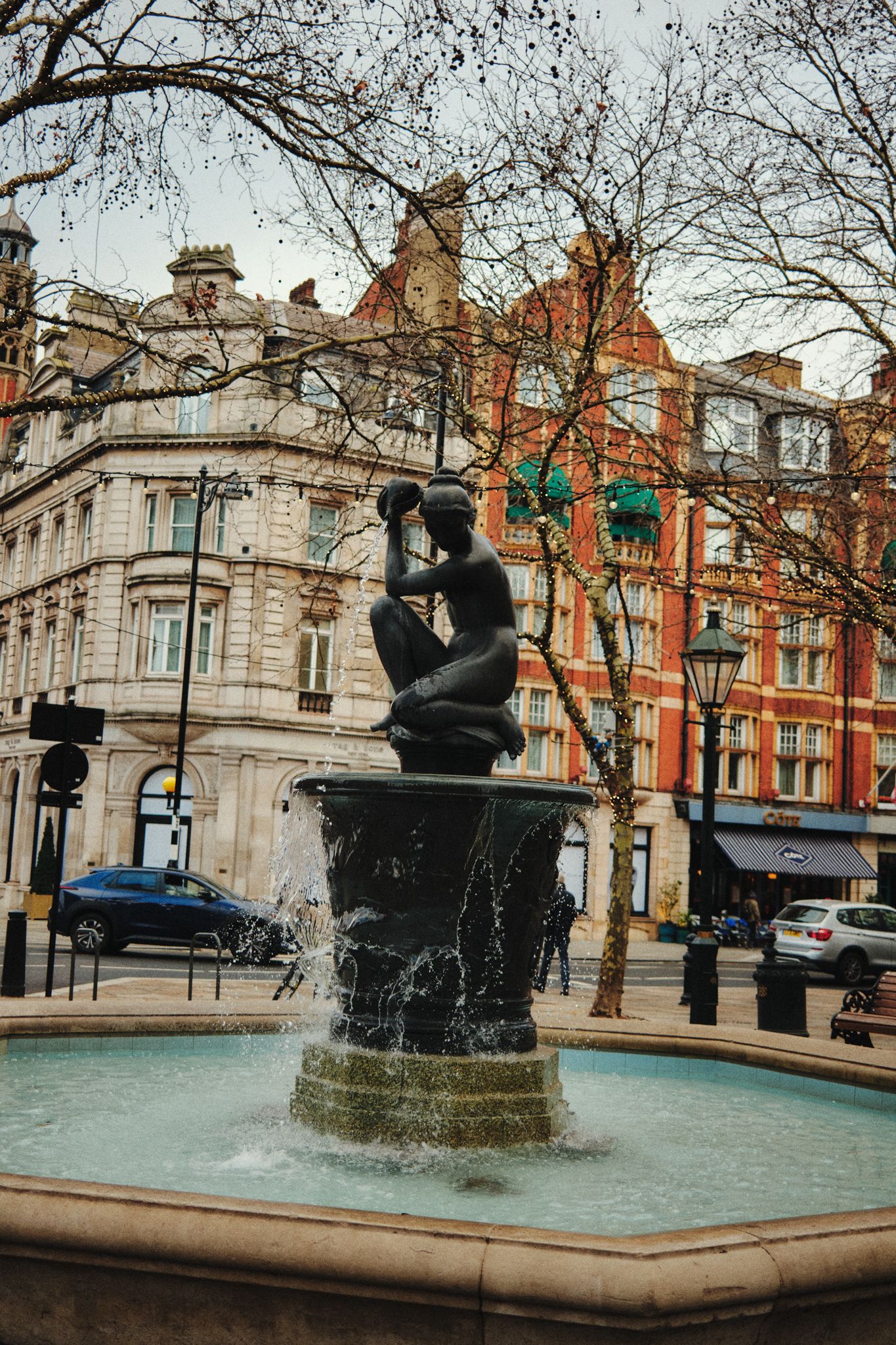A street‑level view of Sloane square in London with a decorative fountain in the foreground featuring a dark sculpture of a seated figure holding a jug. Behind it are leafless trees and elegant, historic multi‑storey buildings with ornate stone and red brick façades, sidewalk cafés, and parked cars. The sky is pale and overcast.