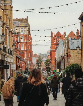 A lively urban street scene with people walking along a pedestrian‑friendly lane flanked by low brick buildings housing shops and cafés. Strings of hanging lights crisscross above the street, and outdoor seating from nearby restaurants lines the sidewalks. The architecture is a mix of warm red and yellow brick, with awnings and signs visible on the storefronts, creating a welcoming neighbourhood atmosphere.