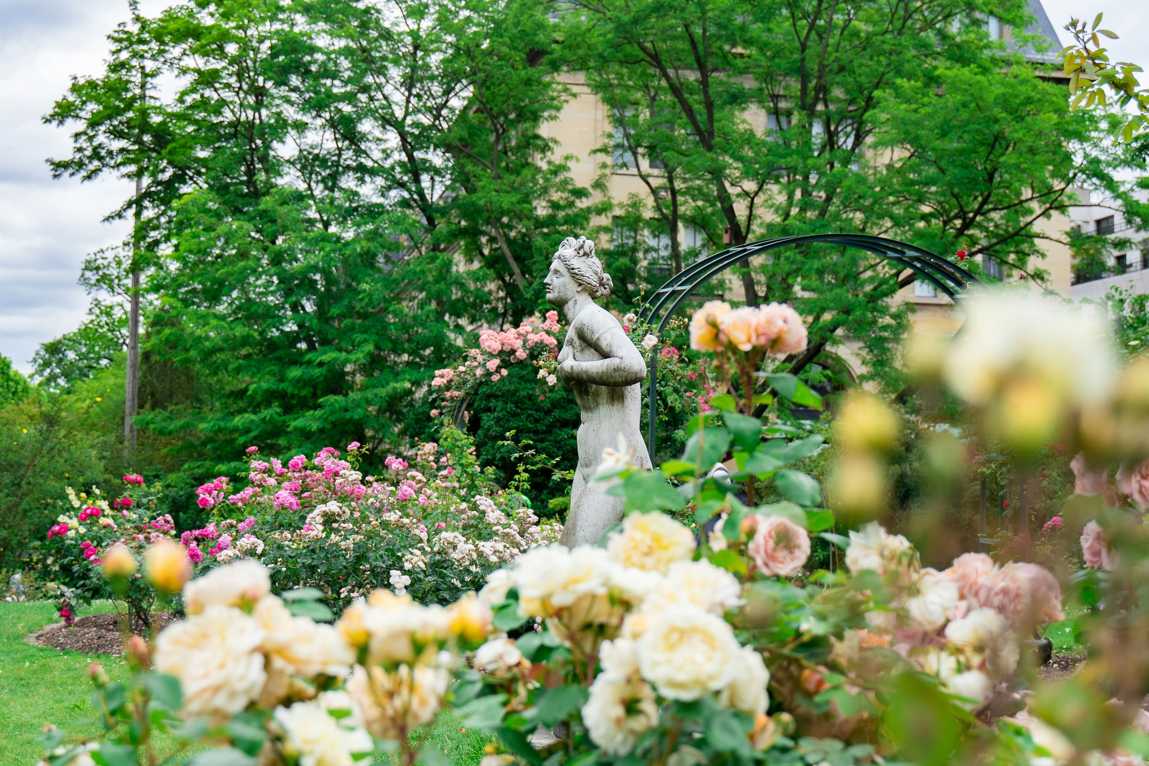 A stone statue of a woman framed by lush greenery and blooming roses in a formal garden setting, with trees and part of a building visible in the background.