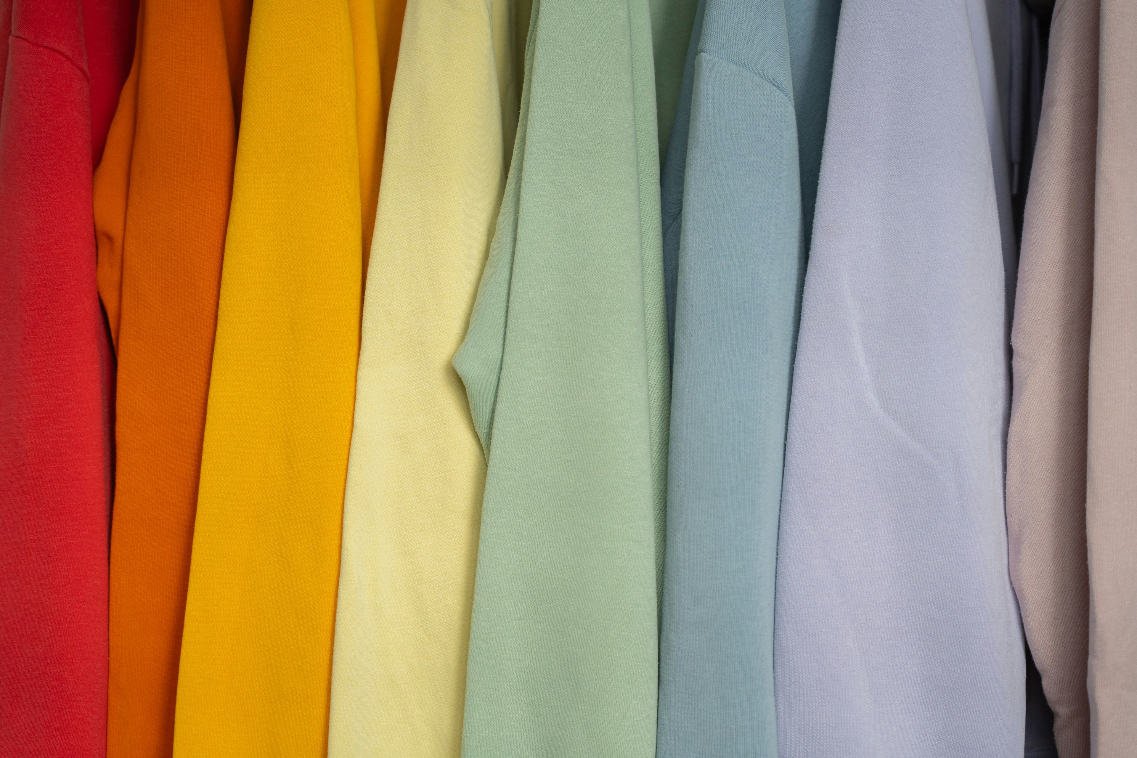 A row of neatly hung shirts or sweatshirts in a range of colors — red, orange, yellow, light green, light blue, and pastel tones — displayed side by side on a clothing rack.