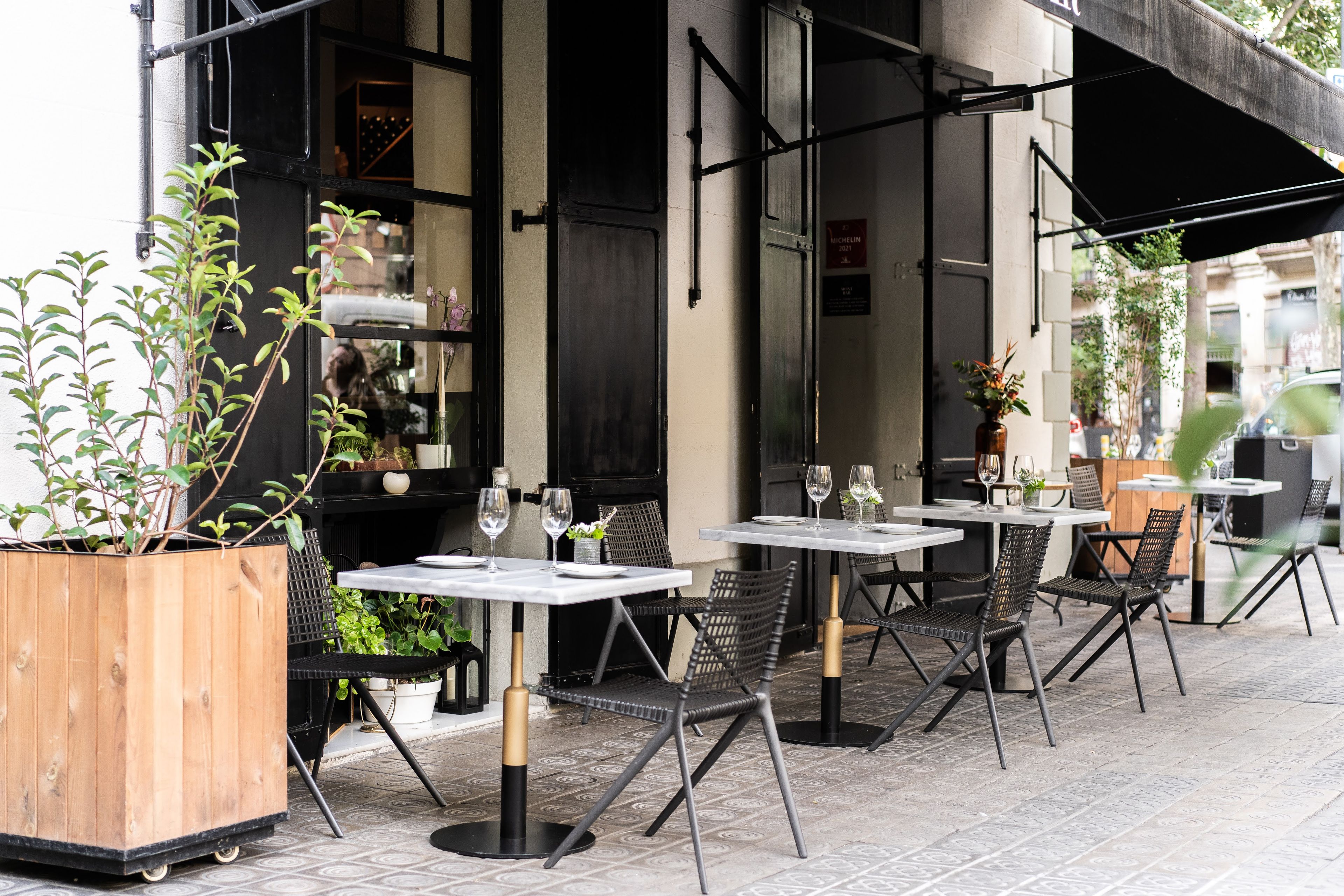 Outdoor terrace seating at Mont Bar in Barcelona with dining tables, chairs, and planters along a city sidewalk.